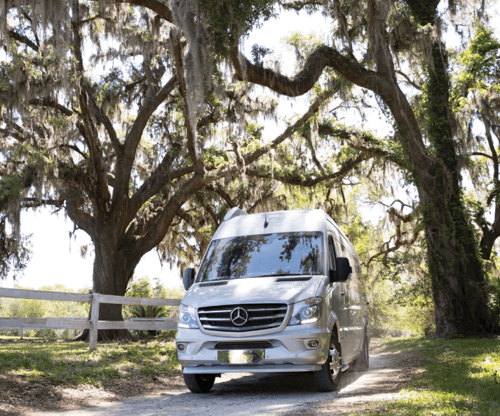 A silver van driving down a rural road with large oak trees overhead.